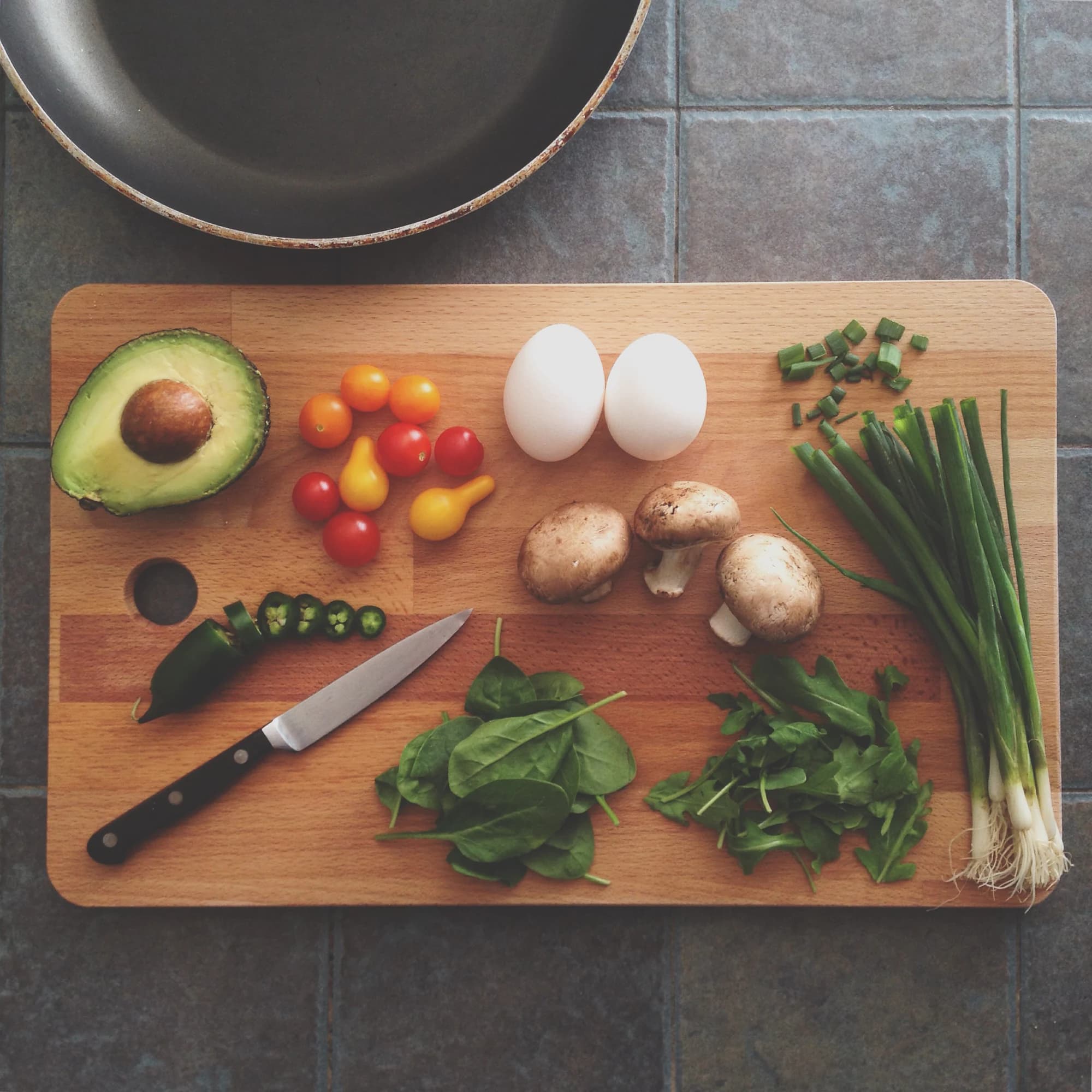 A wooden board laid out with fresh vegetables and eggs beside a pan, in preparation for cooking.