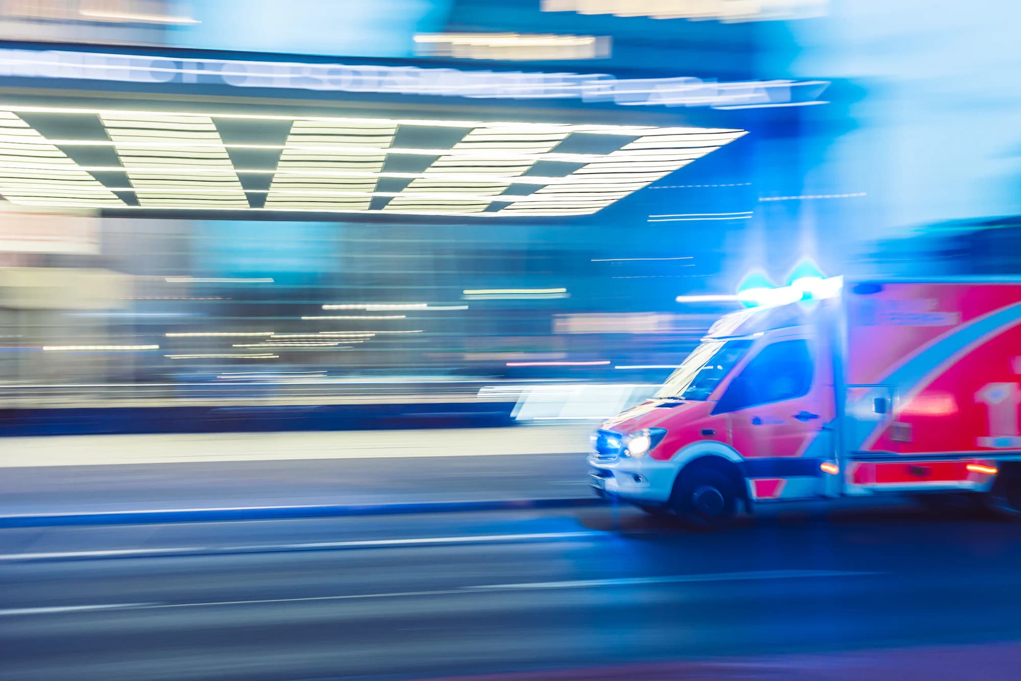 An ambulance moving at speed past a lit hospital entrance, its lights blurred by the exposure.