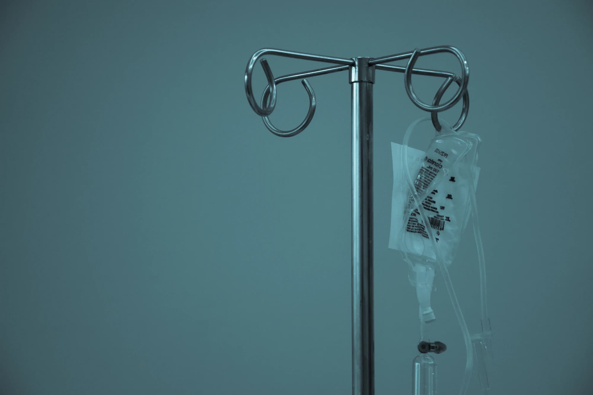 An intravenous drip stand with a fluid bag hanging from it, lit against a dark clinical wall.