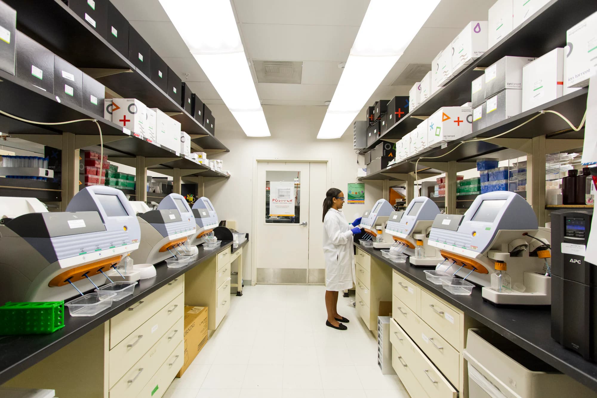 A hospital laboratory bench lined with automated analysers, with a technician working at the far end.