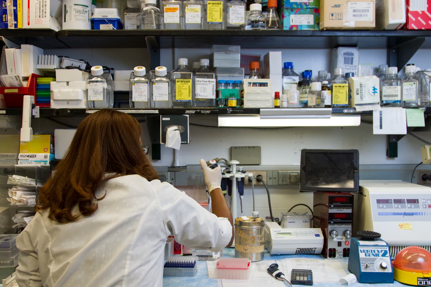 A laboratory bench with reagent shelves and a technician working at an instrument.
