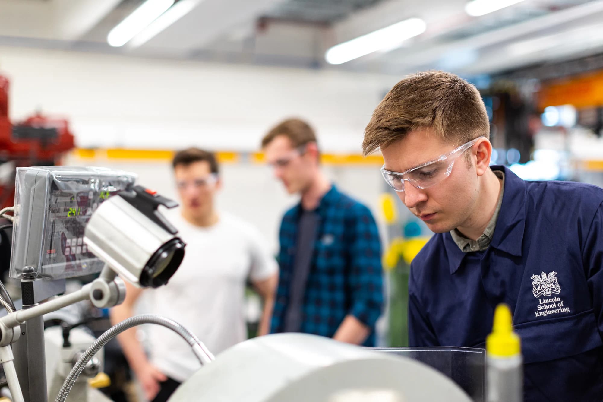 A technician in safety glasses working on equipment at a workshop bench, with colleagues behind.