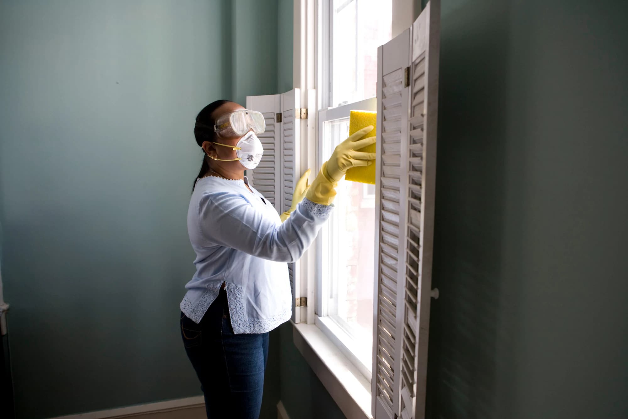 A cleaner in gloves and a mask wiping down a window frame in a bright room.